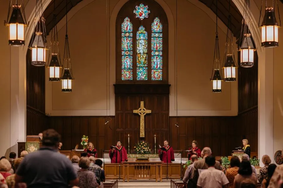 Four people in red robes sing in the wood paneled front apse of SMCC. A carved cross hangs under three stained glass windows behind them.