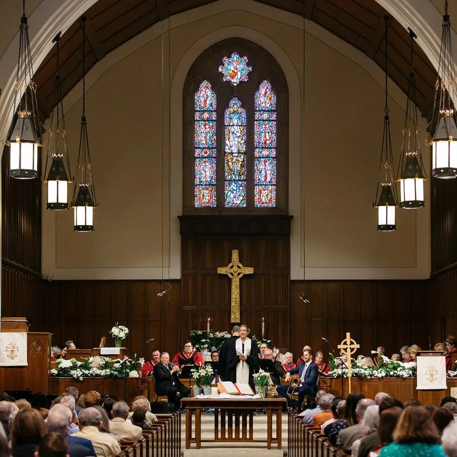 People sit in pews looking toward a man in black robes and a white stole who stands at a table in front of a carved cross and a stained glass window.
