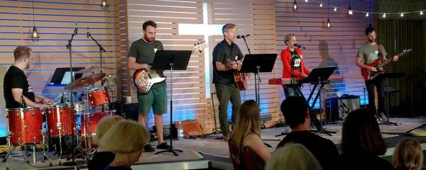 Five members of the Virginia Road Band play electric musical instruments in front of a wood slat wall with a white cross cut out.