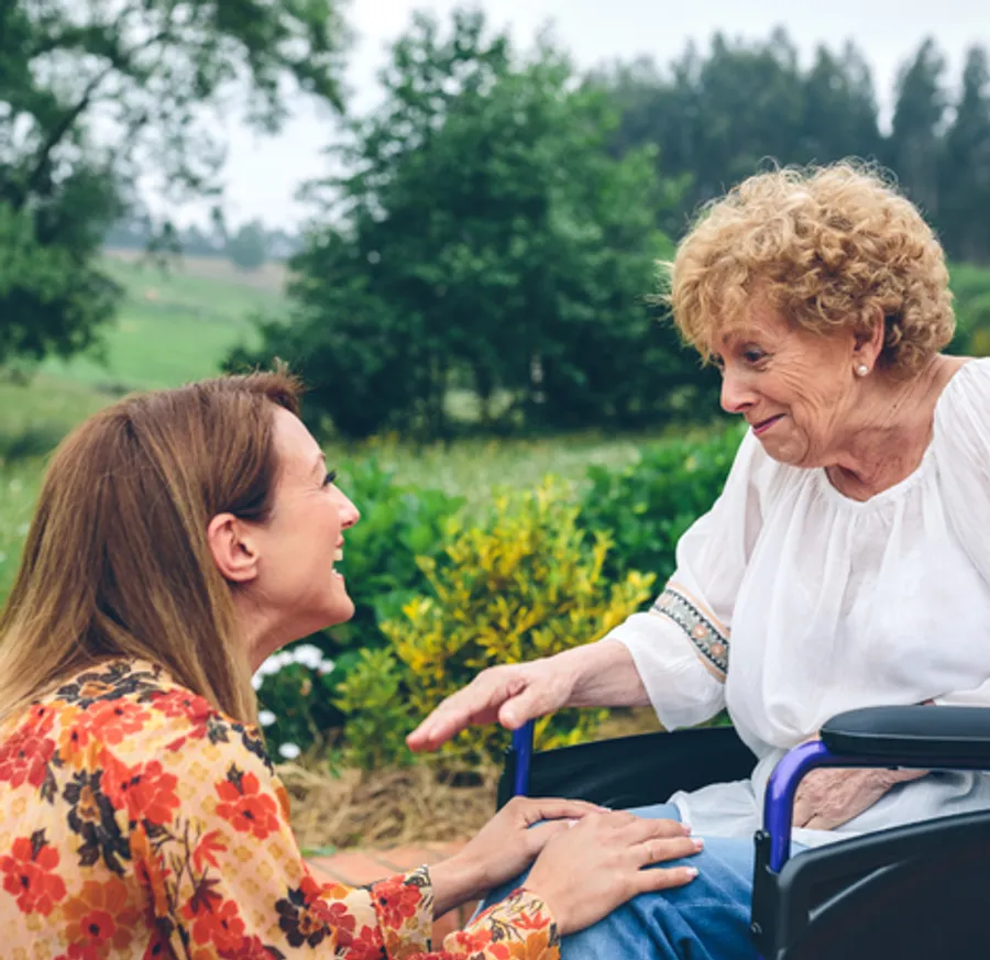 A young woman (left) kneels in front of and older woman (right), sitting in a wheel chair. They smile at one another.