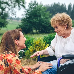 A young woman (left) kneels in front of and older woman (right), sitting in a wheel chair. They smile at one another.
