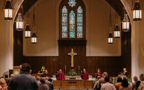 Four singers in red robes sing in the front apse of the SMCC sanctuary. There is an altar, cross, and stained glass window behind them.
