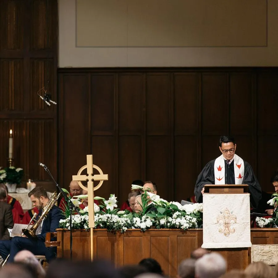 A man wearing black robes and a white stole stands at a wooden lectern in front of lilies and a carved cross hung on a wood paneled wall.