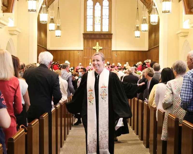 Rev. Jessica Vaughan Lower smiles in black vestments and a white stole while walking from the altar down the center aisle of the sanctuary past pews of parishoners.