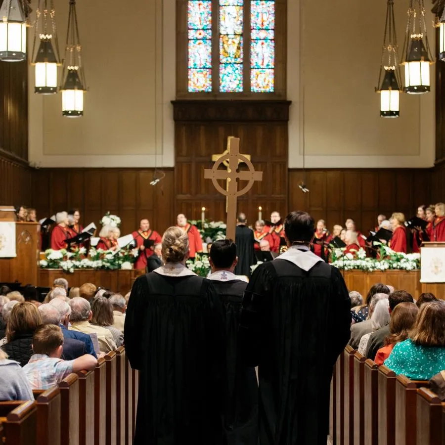 The backs of three people in black vestments and white stoles following a cross down a center aisle past people sitting in pews inside the SMCC sanctuary.