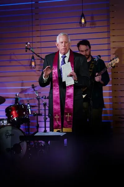Rev. Dr. Tom Tewell wearing a black suit and purple stole with crosses on each end. He preaches in front of a contemporaty worship band.